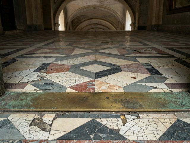 Close-up of a worn brass doorway threshold meeting cracked geometric terrazzo tiles in an old arcade